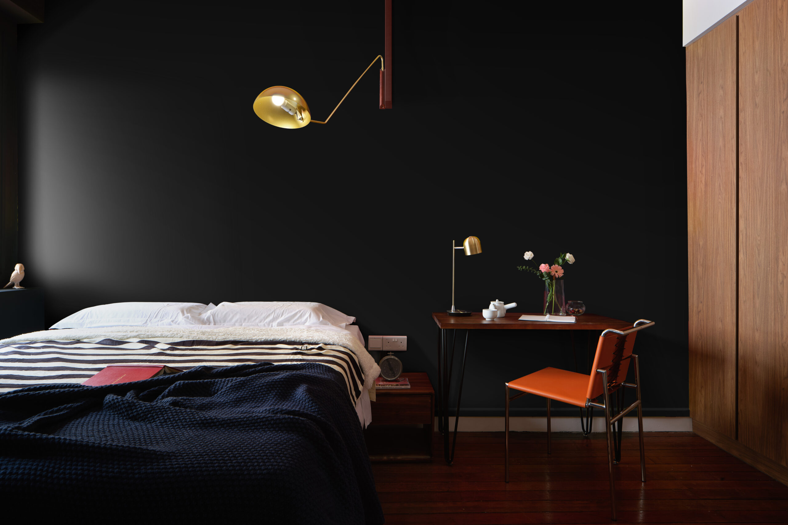 Moody black-walled bedroom in a Chinatown shophouse featuring a low bed, brass wall lamp, wooden desk, and warm minimalist interior styling