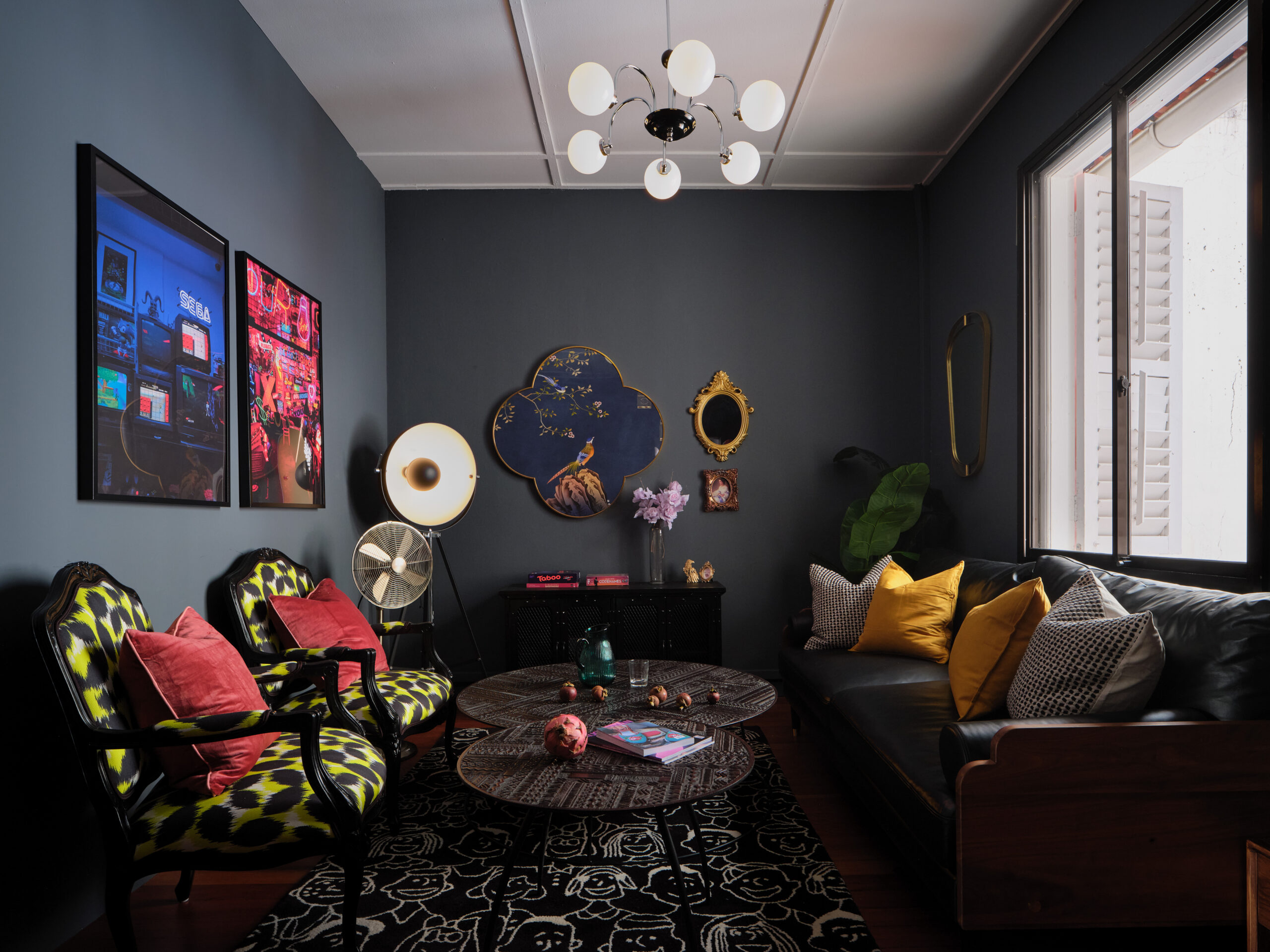 Eclectic living room in a Chinatown apartment with dark blue walls, patterned chairs, vintage decor, layered lighting, and a cozy seating area