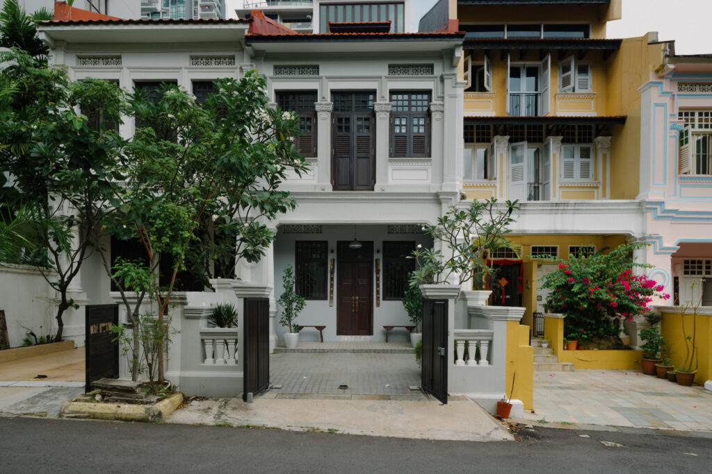 Restored two-storey heritage shophouse with a white facade, dark wooden shutters, and an open black gate, framed by leafy trees on a quiet street.