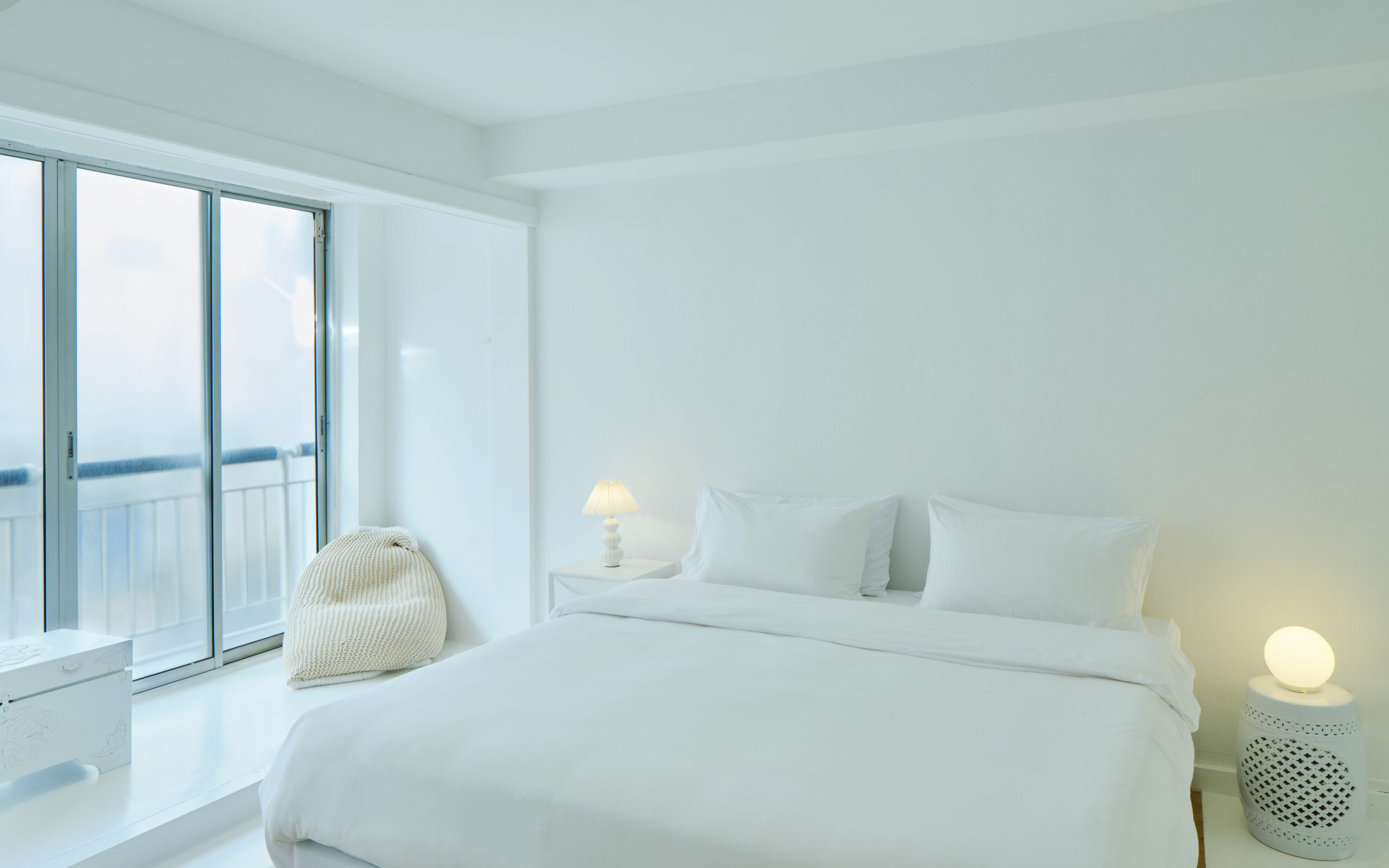 Bright white minimalist bedroom with a neatly made bed, soft lighting, and a large window opening to a balcony.