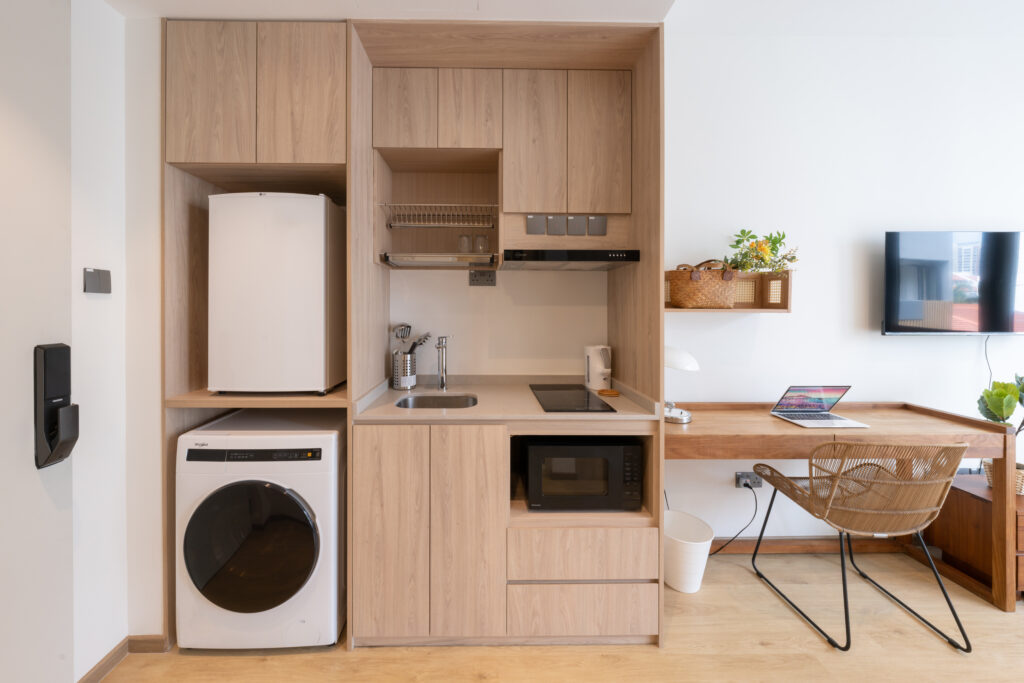 Compact modern kitchenette with light wood cabinetry, built-in fridge, washing machine, microwave, induction cooktop, and sink, next to a minimalist wooden work desk and chair.