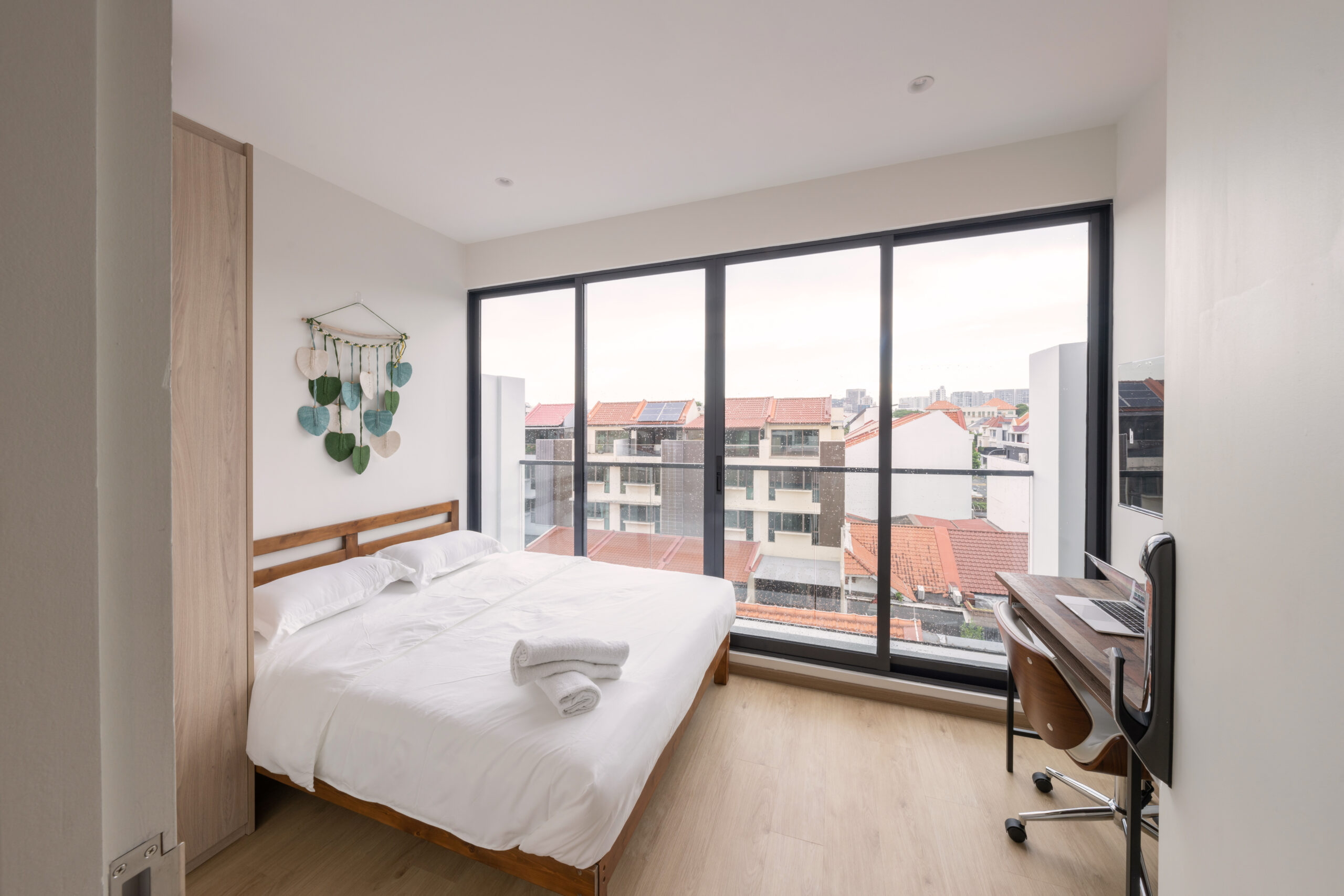 Minimalist bedroom with a wooden bed frame, white bedding and towels, floor-to-ceiling windows overlooking rooftops, and a small work desk by the window.