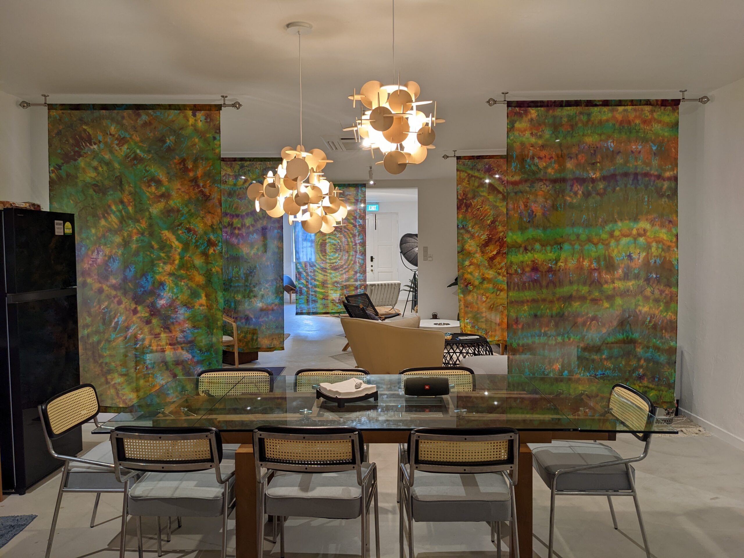 Modern dining area with a glass table, woven chairs, sculptural pendant lights, and colorful patterned curtains dividing the space.