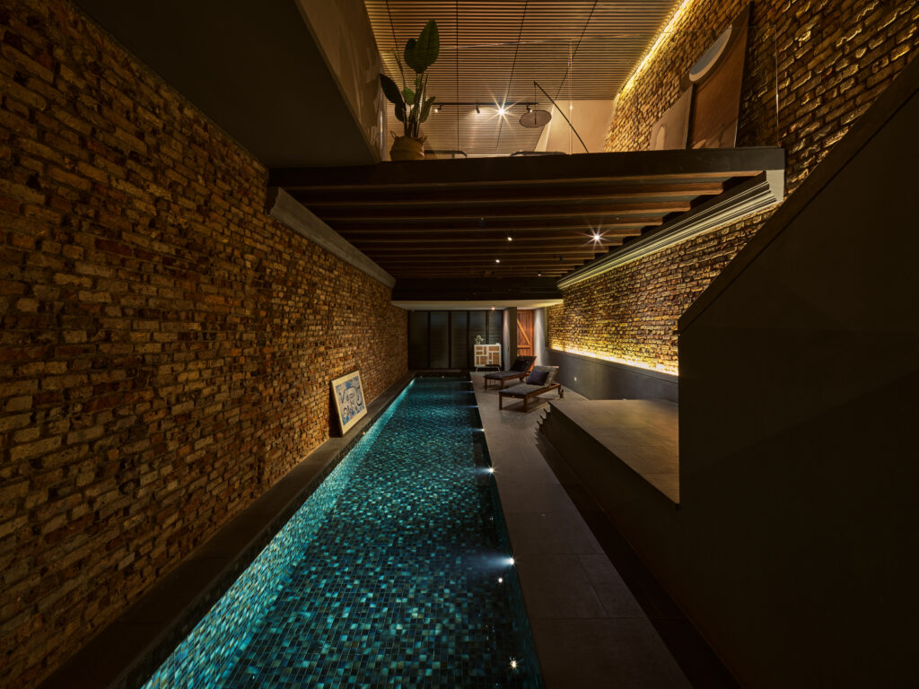Narrow indoor pool with blue mosaic tiles set between exposed brick walls, featuring warm ambient lighting, wooden ceiling slats, and lounge chairs at the far end.