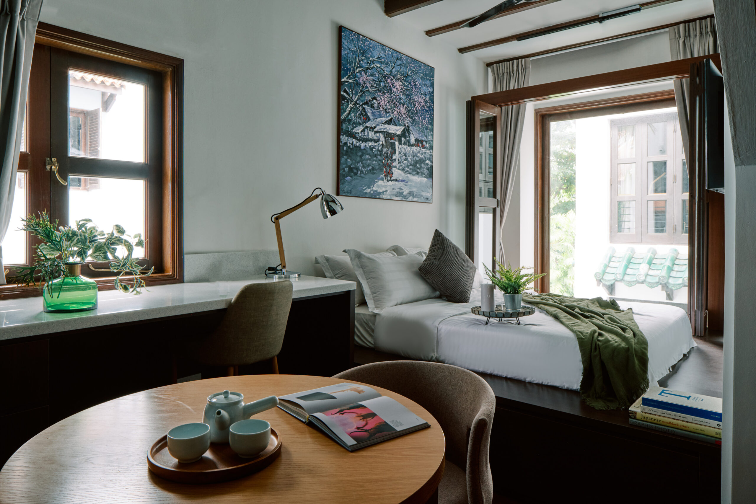 Sunlit bedroom with a low bed, writing desk, and open wooden windows overlooking greenery.