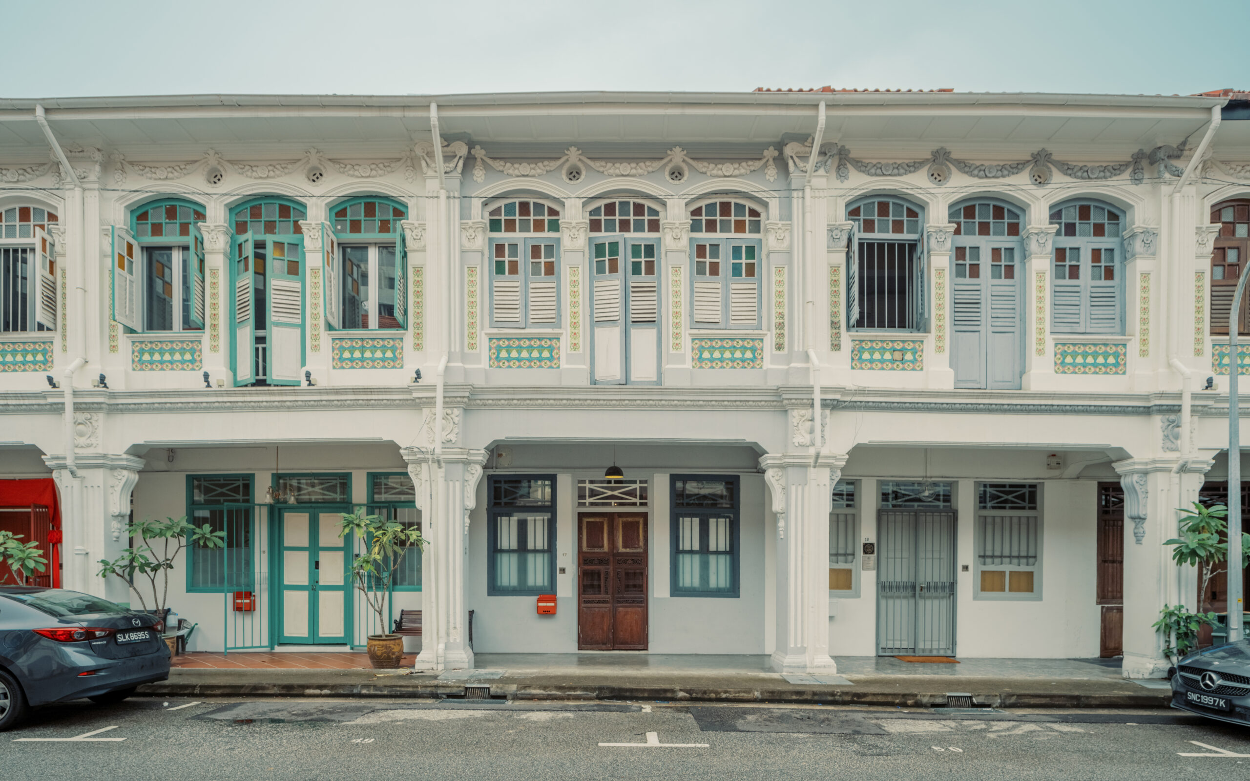 Front view of a traditional shophouse with ornate white facade, pastel blue shutters, and decorative tile panels, photographed from across a quiet street.