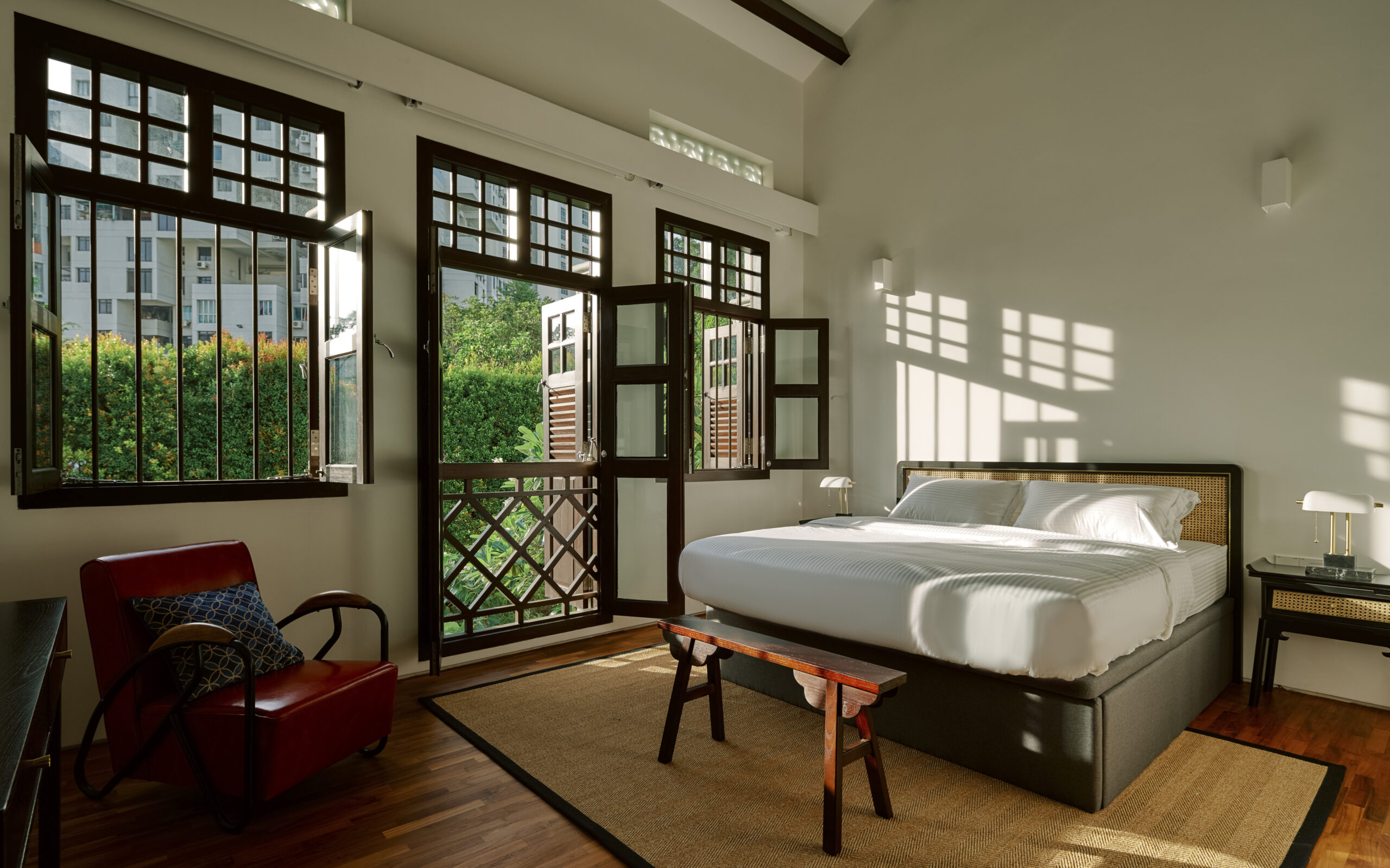 Bright bedroom with a white bed, dark wood furniture, and French doors opening to a small balcony, sunlight casting window shadows on the wall.