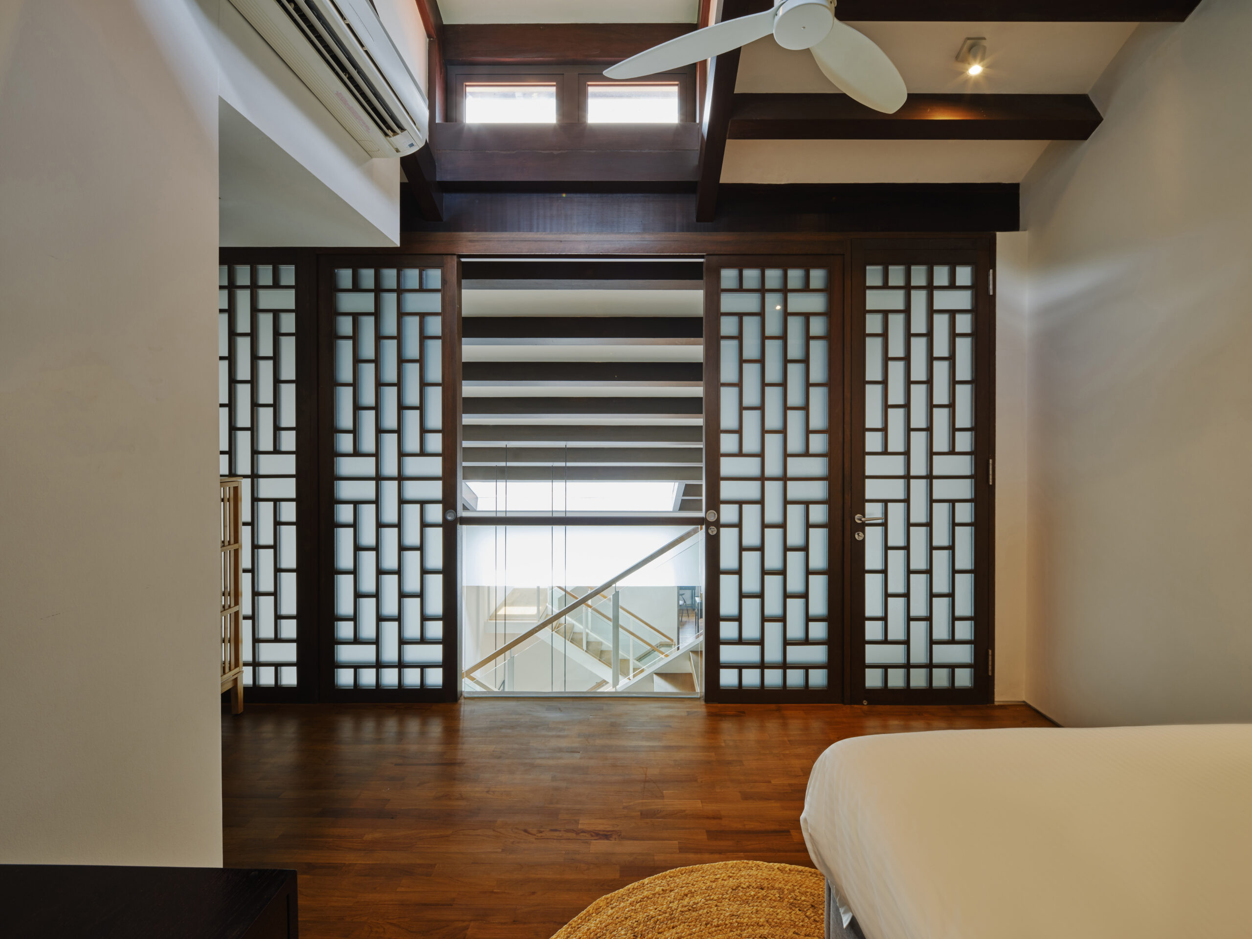 Spacious bedroom with wooden floors, exposed ceiling beams, a ceiling fan, and decorative sliding wooden screens overlooking an interior staircase.