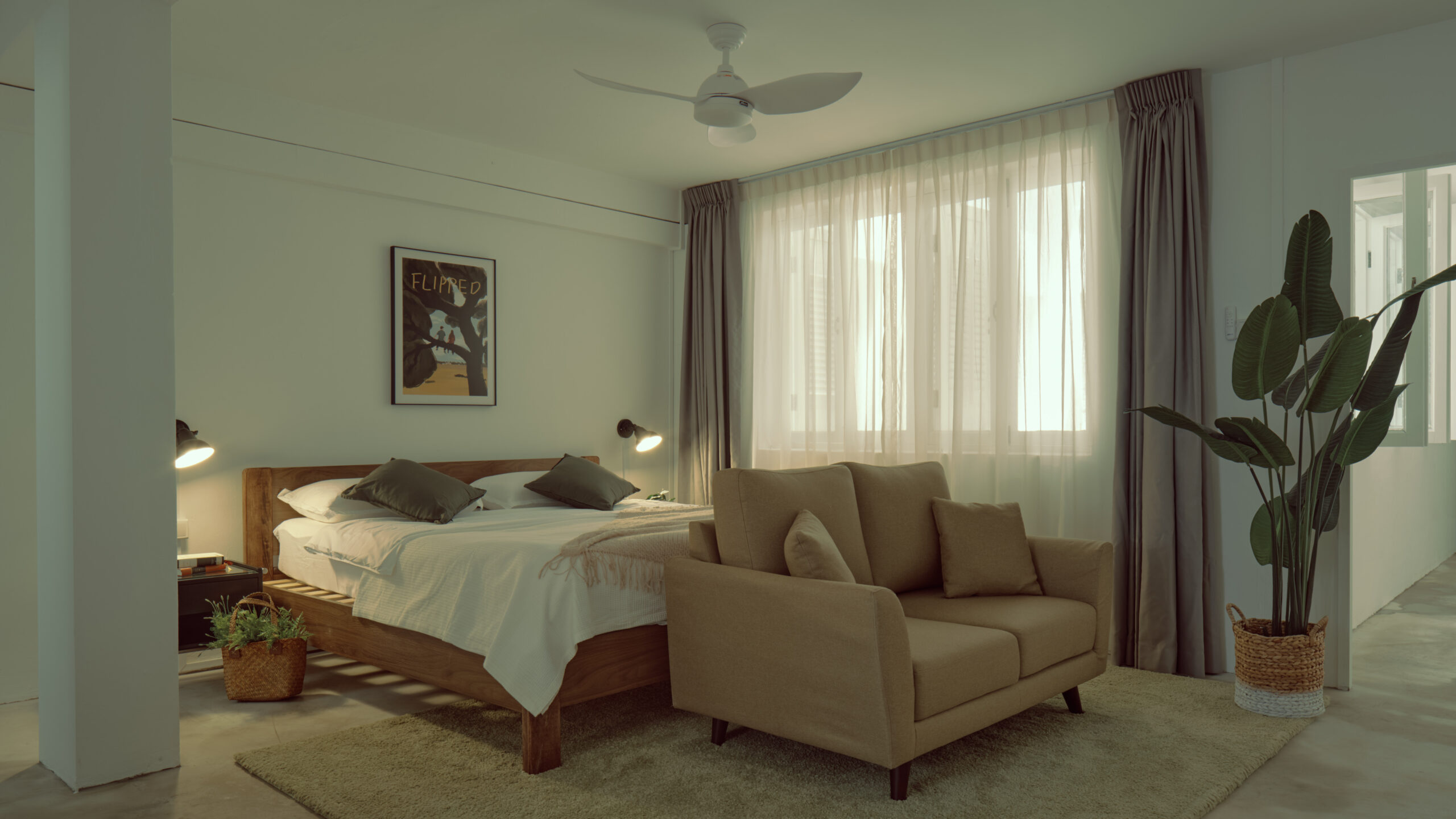 Spacious bedroom with a wooden bed, neutral sofa, soft lighting, sheer curtains, ceiling fan, and indoor plants.